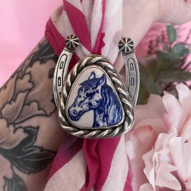 Handmade sterling silver horseshoe bolo tie with a blue-and-white ceramic horse centerpiece, worn on a pink scarf. Sample bolo tie for jewelry making class at The Jeweler's Saw in Austin
