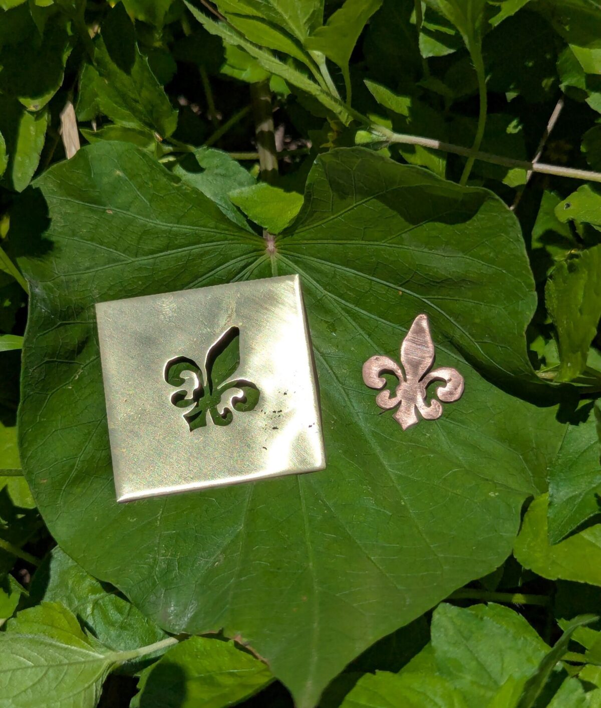 Hand-cut fleur-de-lis jewelry piece and copper cutout on a green leaf, made in a metalsmithing class at The Jeweler’s Saw in Austin.